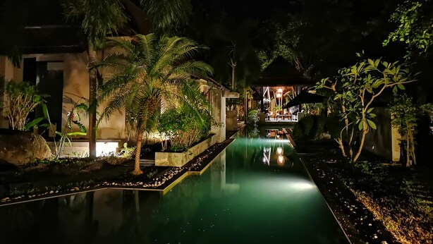 A nighttime view of a pool area with a reflection, surrounded by palm trees and a building with a covered seating area. - Montra Samui (Koh Samui)