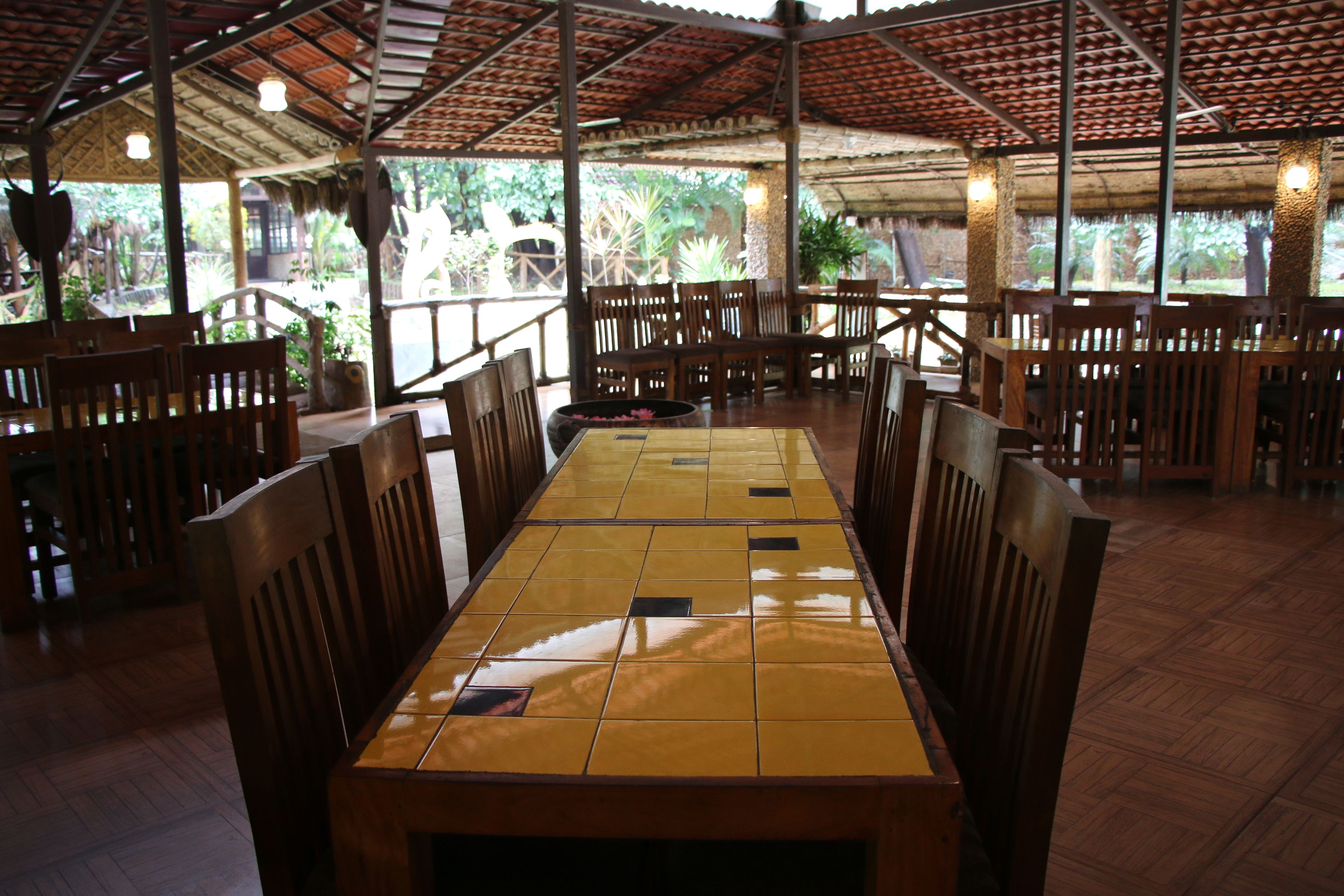 A restaurant with a long wooden table and chairs, a tiled floor, and a thatched roof.