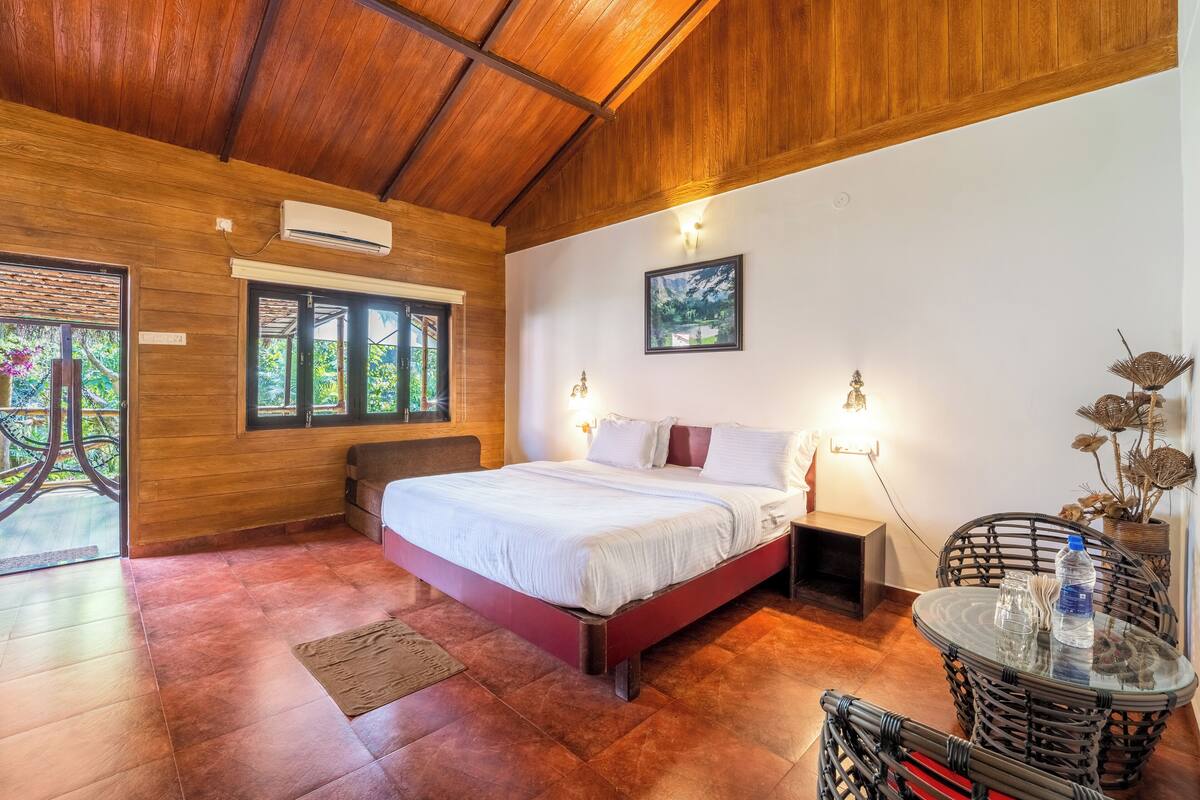 A bedroom with a wooden ceiling, a bed with white bedding, a small table with a water bottle, and a wicker chair.