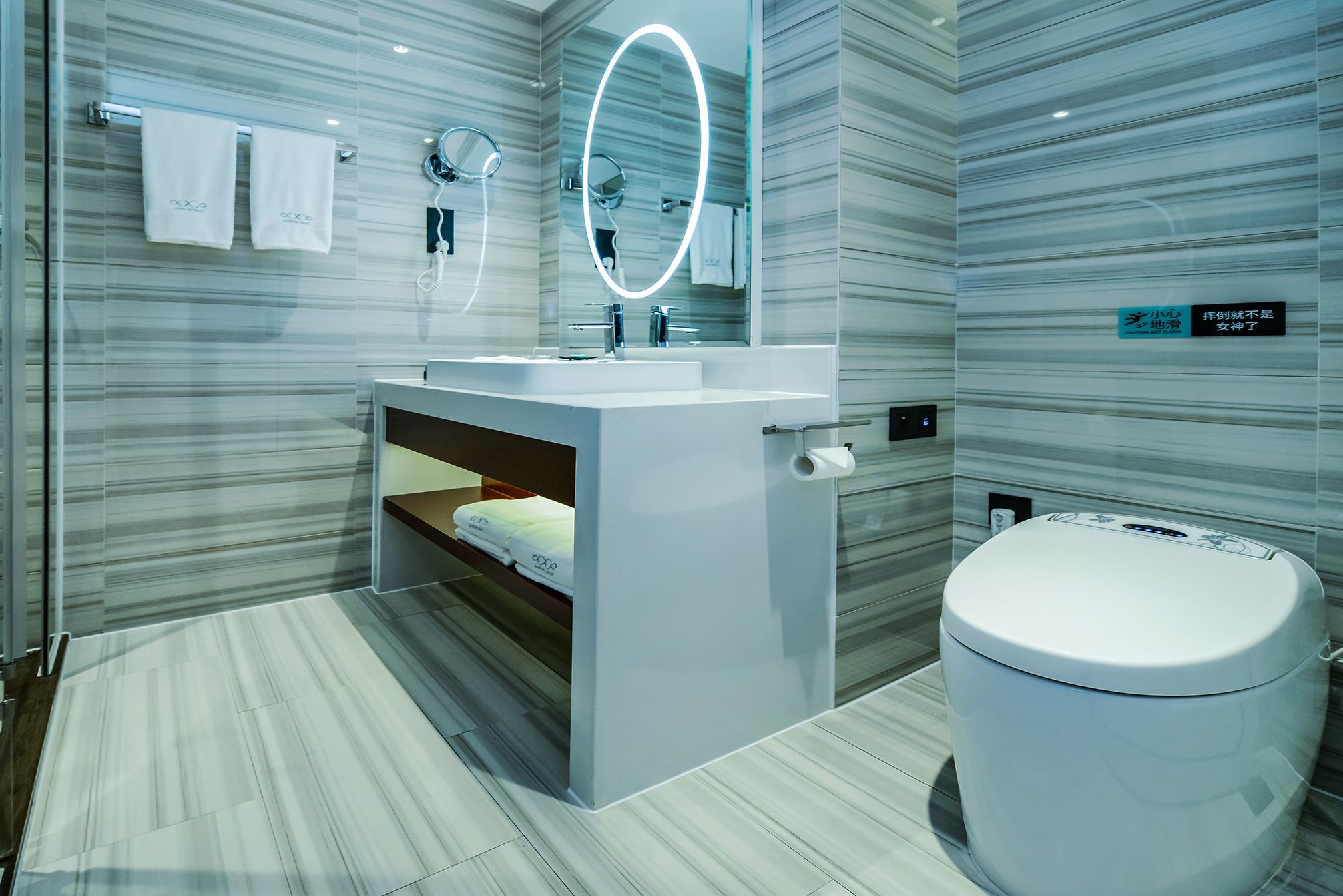 A modern bathroom with a marble wall, a white sink, a mirror with a light, and a toilet.