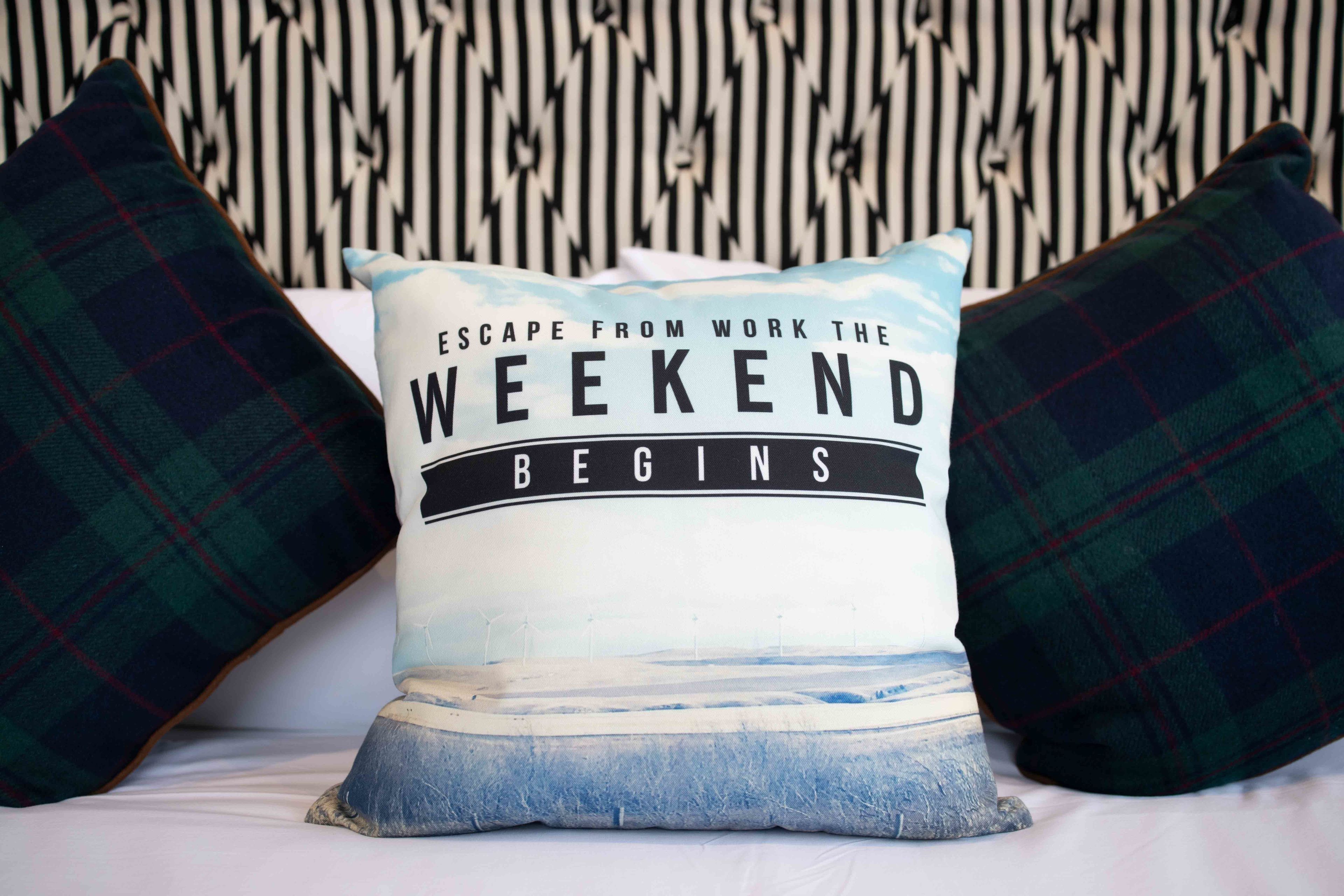 A pillow with the text "Escape from work the weekend begins" on a bed with plaid pillows and a patterned wall.