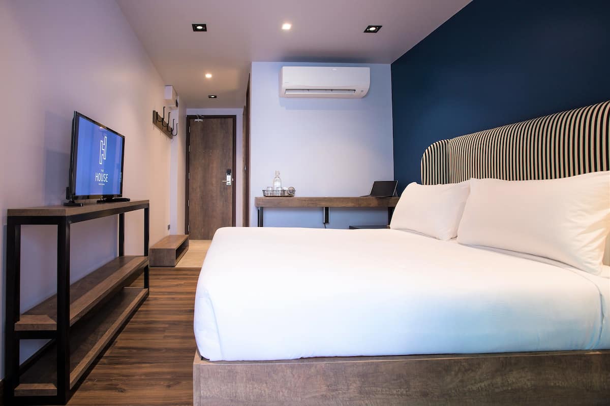 Una habitación de hotel moderna con una cama grande, un televisor sobre una consola de madera, un escritorio con una computadora portátil y un equipo de aire acondicionado en la pared.