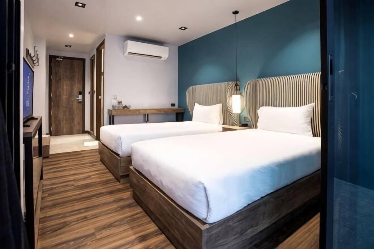 Habitación de hotel con dos camas, suelo de madera, un escritorio con silla, aire acondicionado de pared y una lámpara colgante.