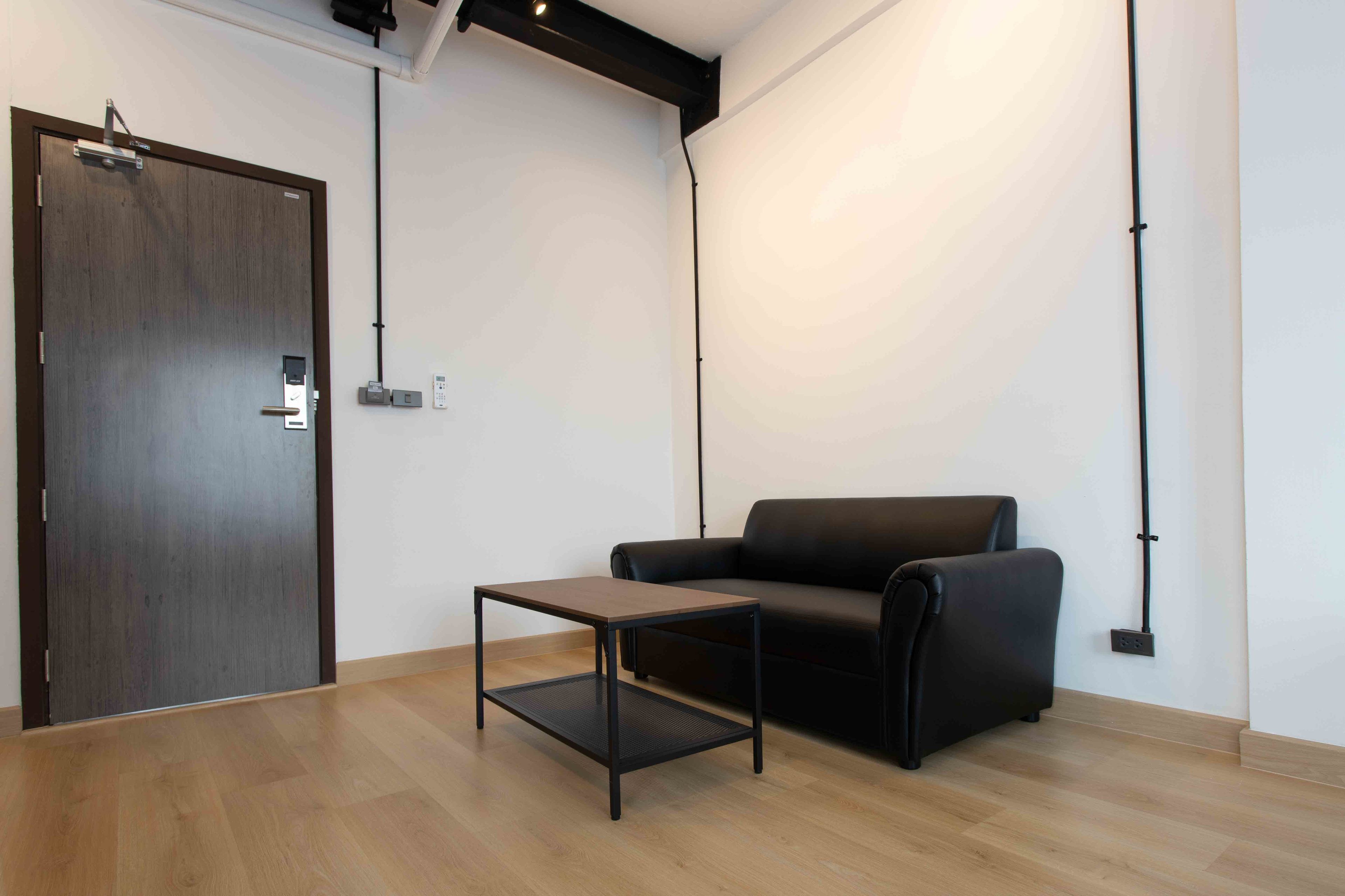A room with a wooden floor, a black leather sofa, a small black table, a wooden door, and a white wall with a black pipe running vertically.