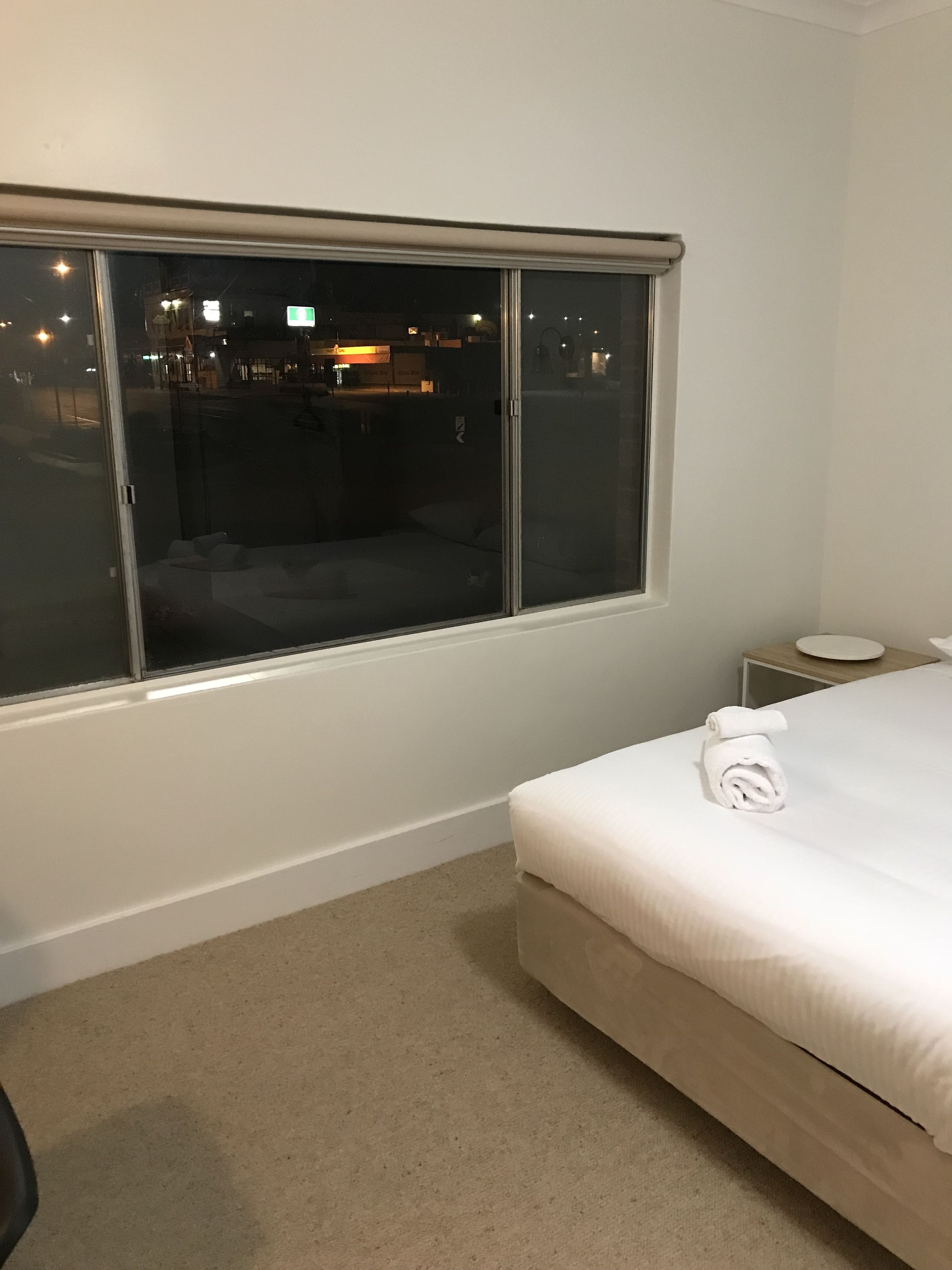A hotel room with a large window, a bed with a rolled towel, a small bedside table, and a carpeted floor.
