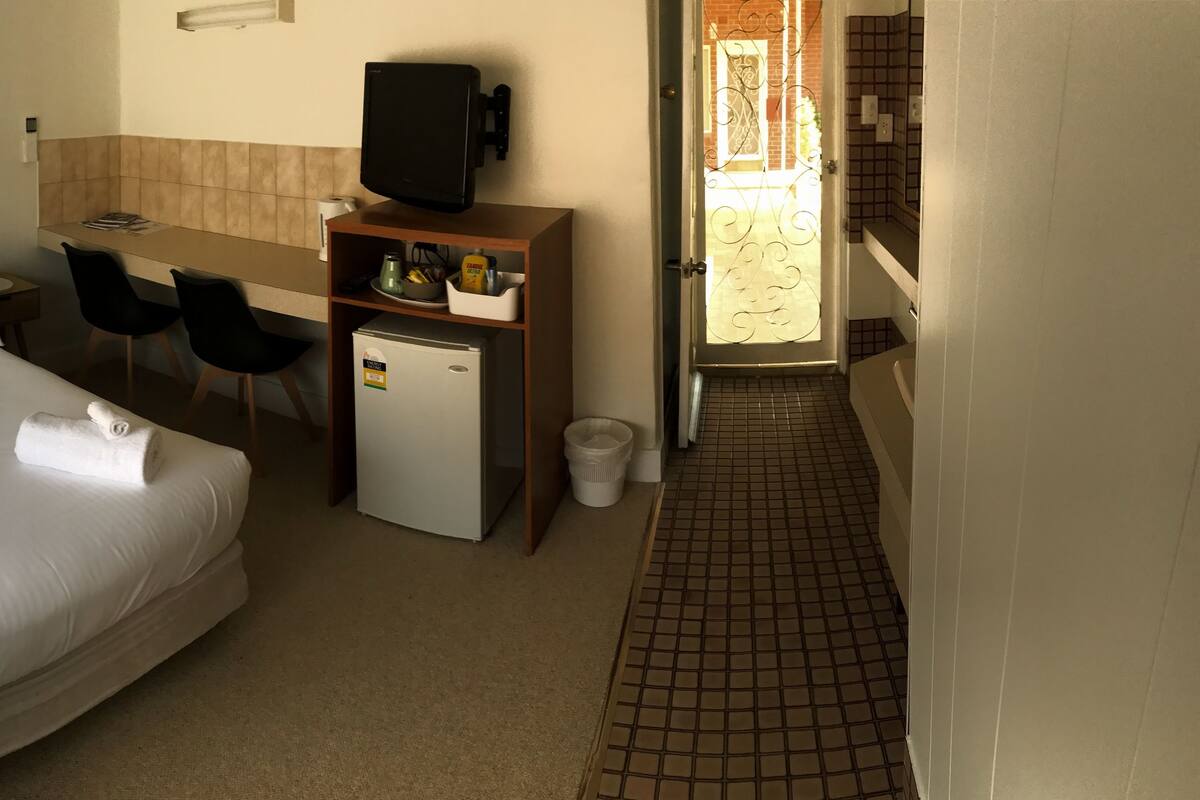 A hotel room with a bed, a desk, a chair, a small refrigerator, and a bathroom visible through an open door.