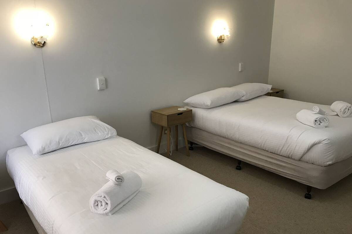 A hotel room with two single beds, each with a rolled towel, a wooden nightstand, and wall-mounted lights.