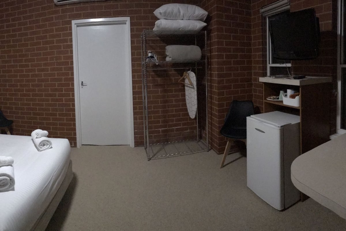 A small, compact hotel room with a single bed, a desk, a chair, a small table, a wardrobe, a TV, and a refrigerator.