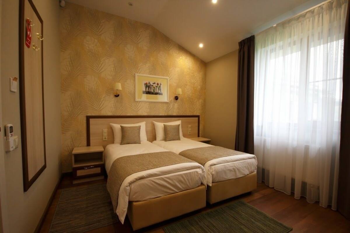 Habitación de hotel con cama doble, mesita de noche pequeña, un cuadro en la pared y una ventana con cortinas traslúcidas.