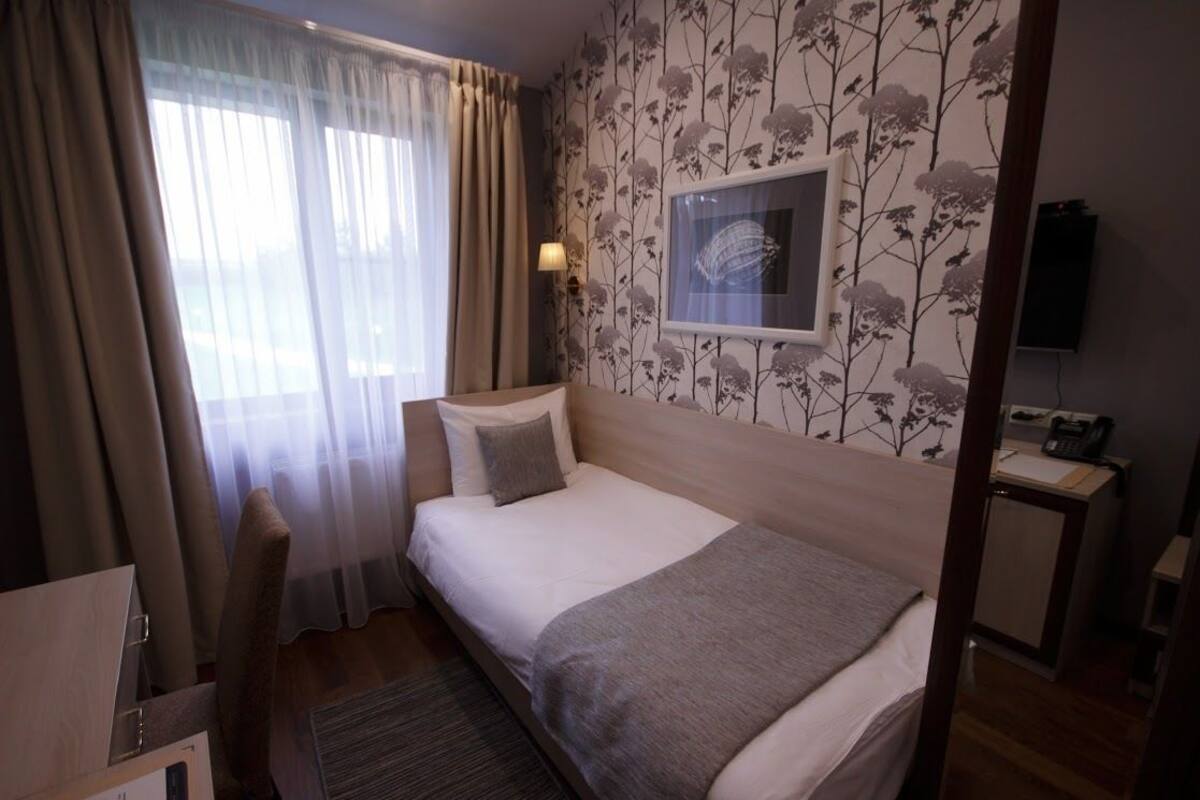 Una habitación de hotel con una cama individual, un escritorio con silla, una mesita pequeña, un teléfono y un televisor instalado en la pared.