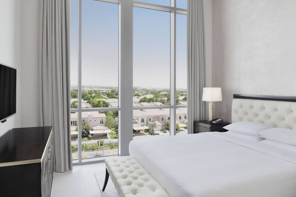 Ein Hotelzimmer mit großem Fenster und Blick auf die Stadt, einem Bett mit weißer Bettwäsche, einem Nachttisch mit Lampe sowie einem Fernseher auf der linken Seite.