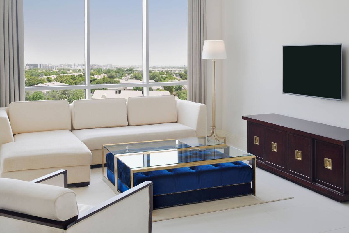 Ein modernes Wohnzimmer mit einer Couch, einem Glastisch, einem blauen Pouf, einer dunkelholznen Kommode und einem an der Wand befestigten Flachbildfernseher.