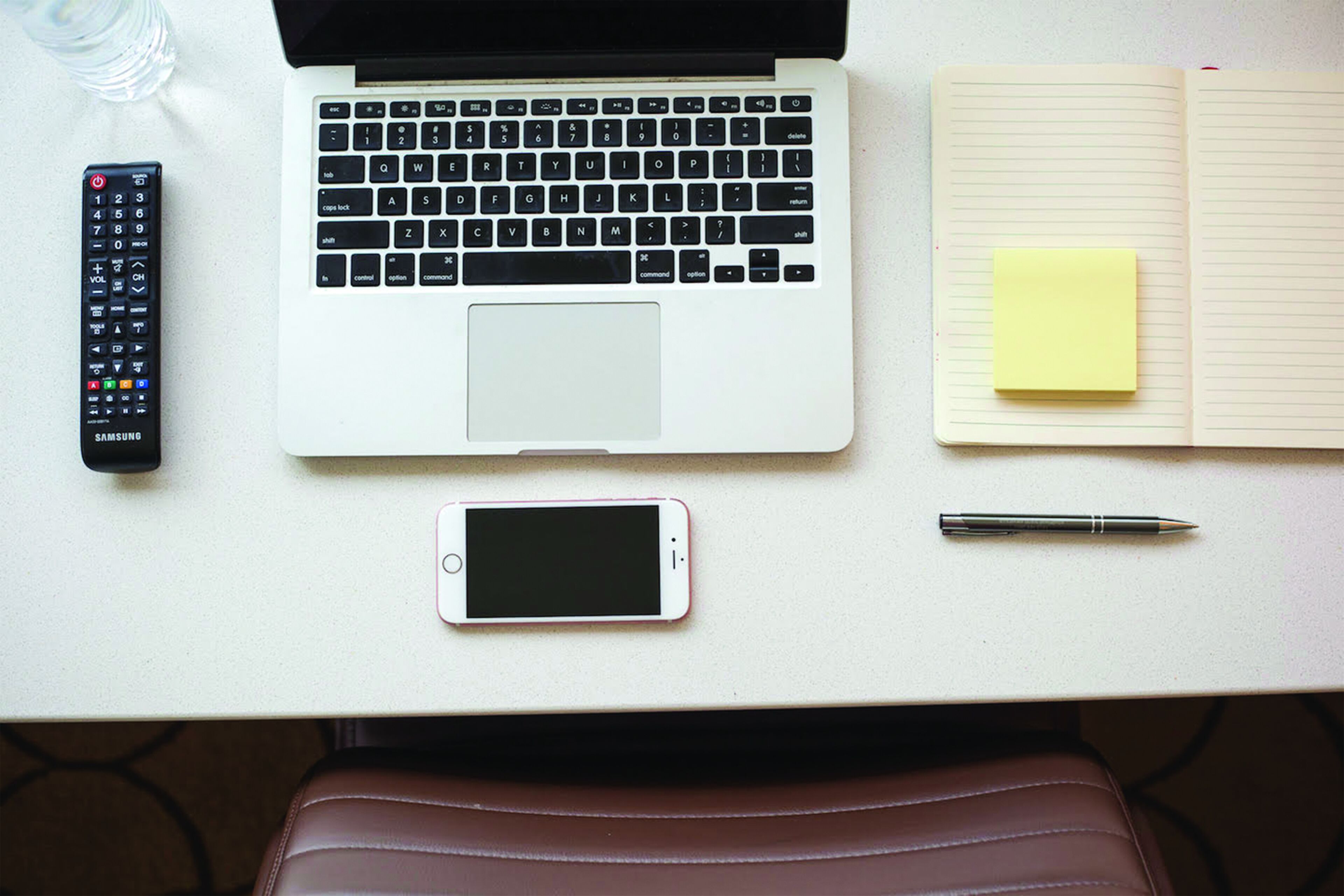 A laptop, smartphone, remote control, pen, and a notebook with a sticky note on a white table.