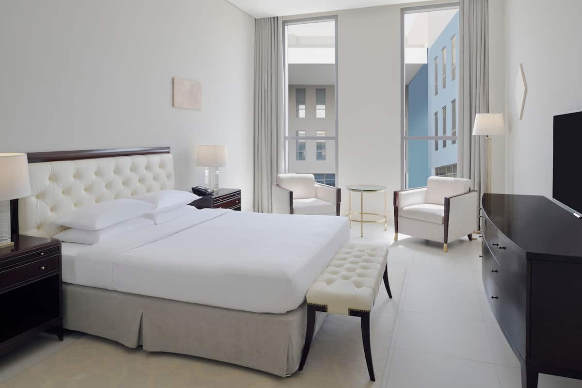 Ein Hotelzimmer mit einem großen Bett, einem Schreibtisch, einem Sessel, einem Fernseher und Blick auf Gebäude durch das Fenster.