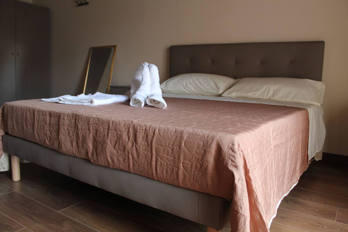 Un lit bien fait, recouvert d’une couverture rose, une pile de serviettes blanches pliées et un miroir accroché au mur.