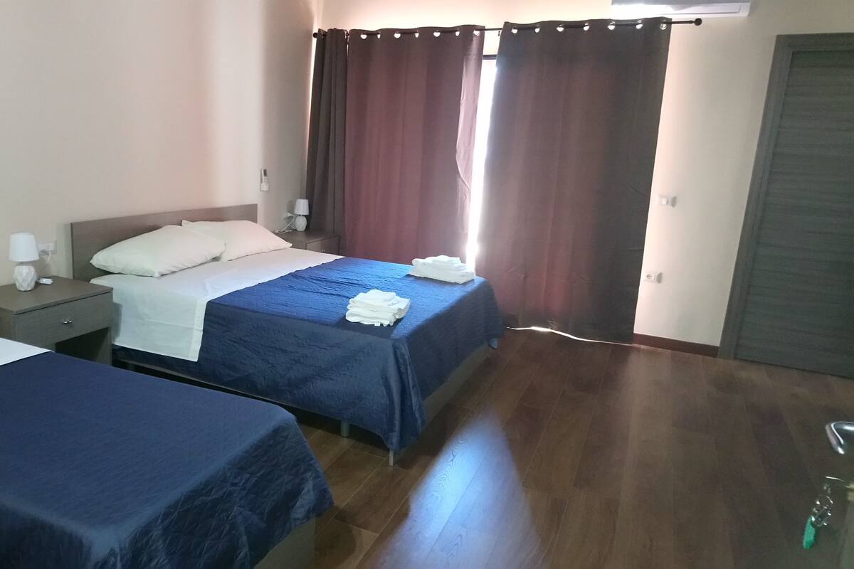 Une chambre d’hôtel avec deux lits, chacun recouvert d’une couvre-lit bleue et agrémenté d’oreillers blancs. De chaque côté des lits, il y a une table de chevet avec une lampe et un téléphone. La chambre est dotée d’un plancher en bois et la porte se trouve sur le côté droit.