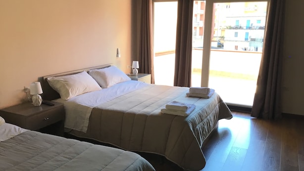 Een hotelkamer met twee bedden, een nachtkastje met een lamp en uitzicht op de straat door grote ramen.