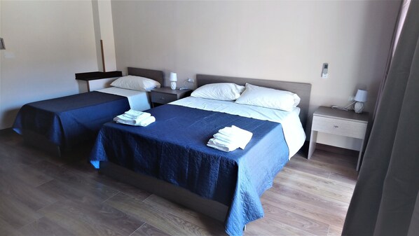 Une chambre d’hôtel avec deux lits, chacun recouvert d’une couverture bleue et agrémenté d’oreillers blancs. Il y a deux tables de chevet, l’une avec une lampe et l’autre avec un téléphone. La chambre a un plancher en bois et un mur uni.