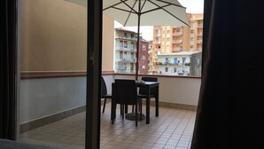 Een balkon met een tafel en stoelen, een grote parasol en uitzicht op meerlaagsgebouwen.