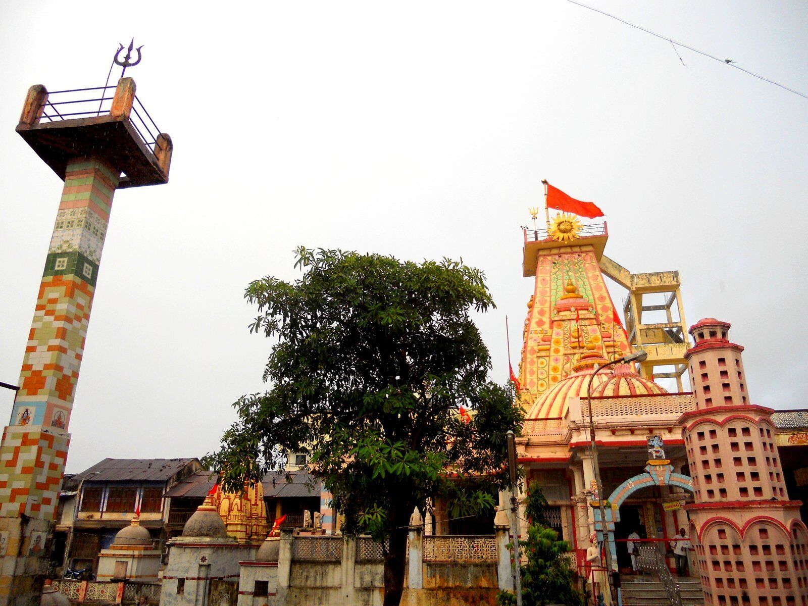 A temple with a large tree in front, a tall pillar with a trident, and a colorful structure with a flag.