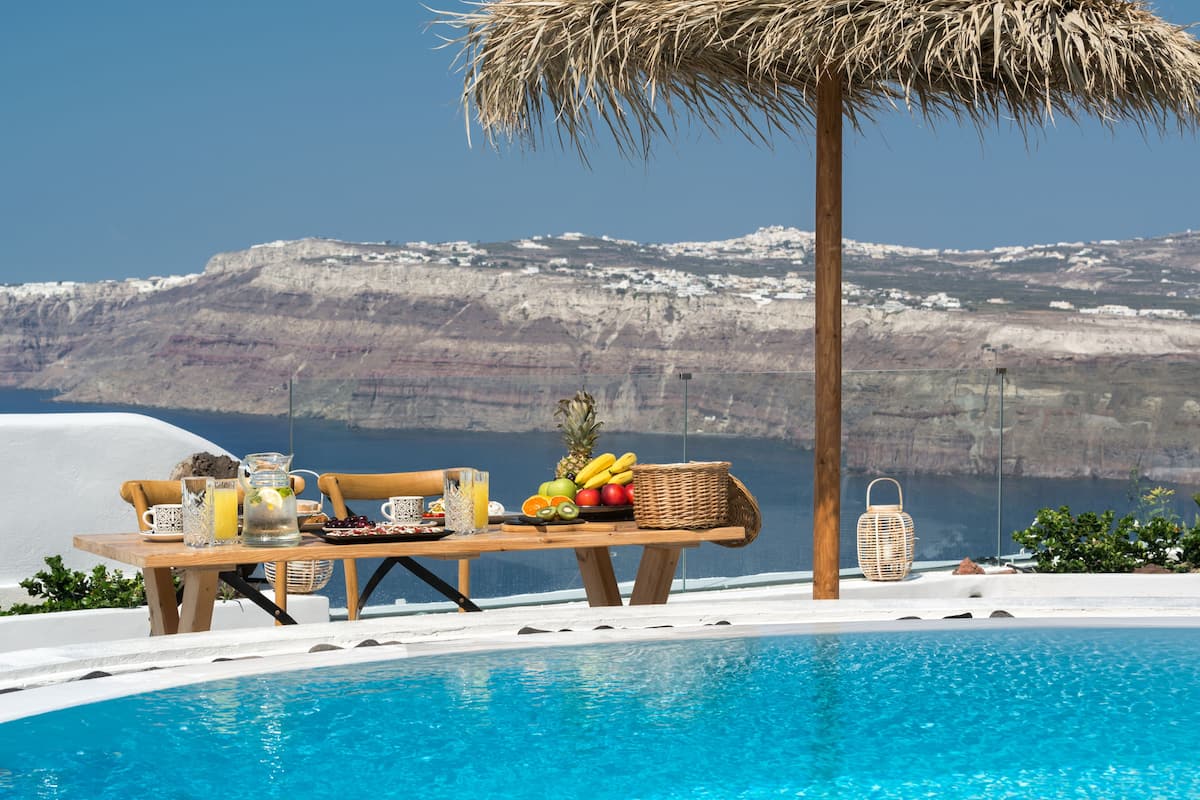 A poolside breakfast setup with a table, fruits, and drinks, overlooking a scenic cliff and ocean.