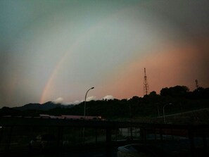 Un arc-en-ciel se dessine au-dessus d’un paysage montagneux, avec une route et des bâtiments au premier plan.