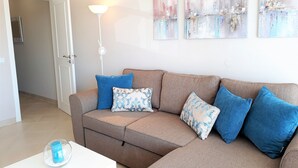 Ein Wohnzimmer mit einem beigefarbenen Ecksofa, blauen und gemusterten Kissen, einem weißen Beistelltisch mit einer blauen Kerze und abstrakter Wandkunst.