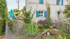 Un bâtiment à deux étages, doté d’une façade blanche et de volets bleus, entouré d’un jardin où poussent différentes espèces de plantes et délimité par une clôture en métal.