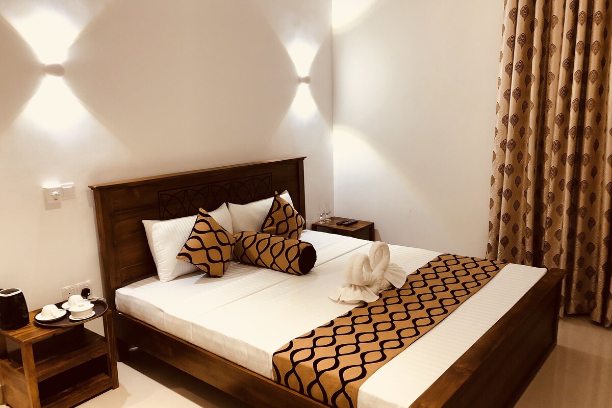 Cama bem arrumada com almofadas estampadas, mesa de cabeceira com bandeja de xícaras e uma cafeteira, e cortina com padrão de folhas.