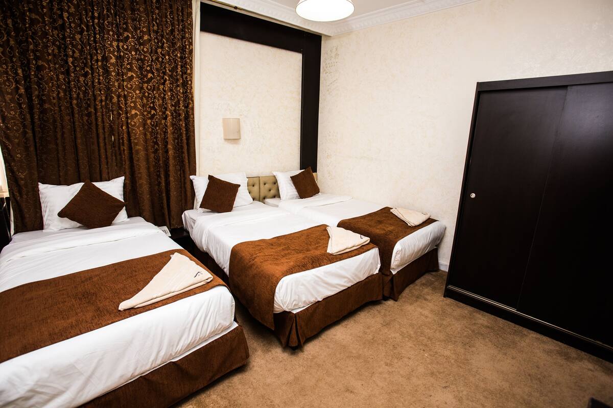 A hotel room with two beds, brown and white bedding, a large window with curtains, a wall-mounted light, and a dark door.