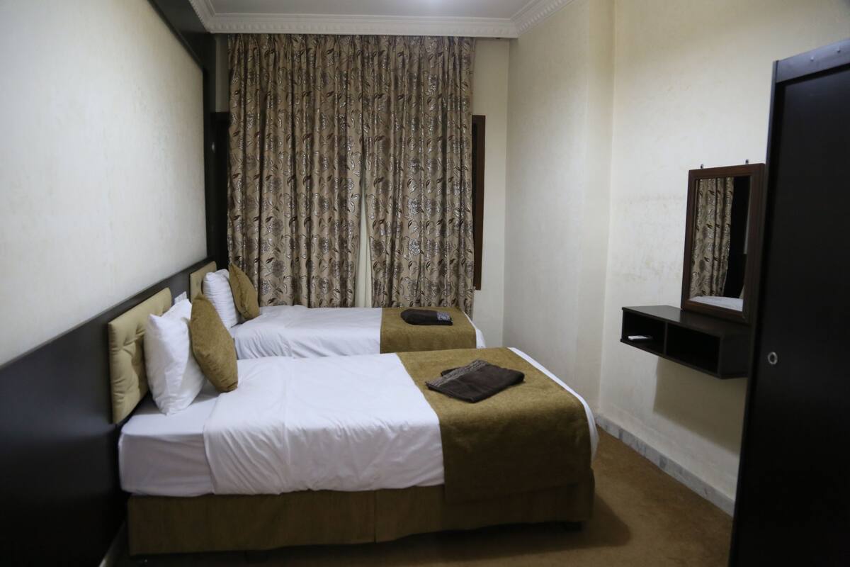 A hotel room with two beds, a dark headboard, a large window with patterned curtains, a small shelf with a mirror, and a carpeted floor.