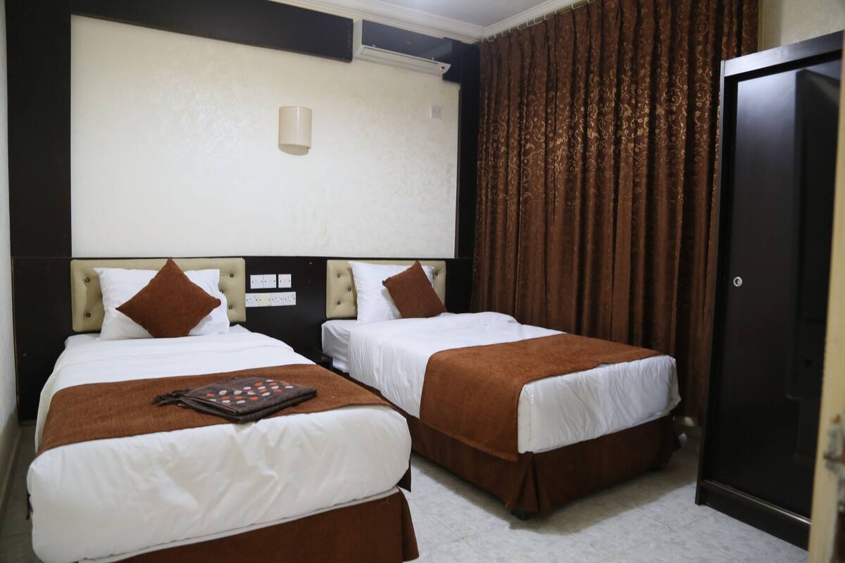 A hotel room with two beds, each with a brown headboard and white bedding. There is a brown throw blanket on one bed and a small brown bag on the other. The room has a dark door and a patterned curtain.