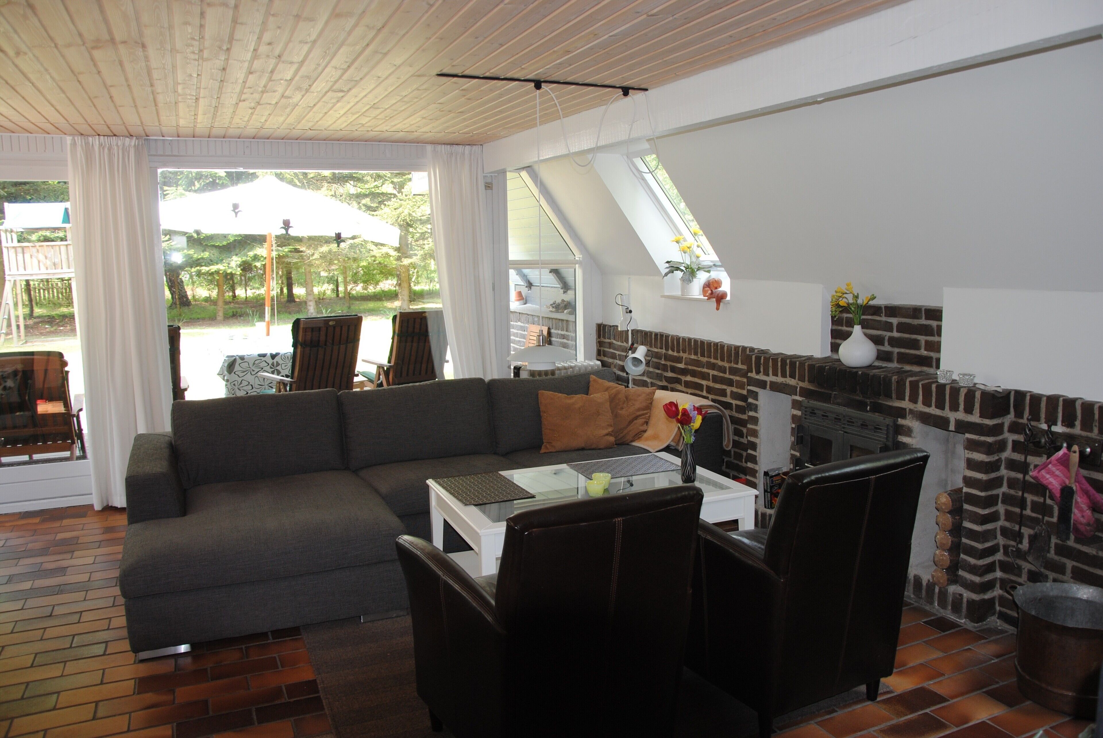 Ein modernes Wohnzimmer mit einem Ecksofa, einem Esstisch mit Stühlen, einem Kamin aus Ziegelsteinen und großen Fenstern mit Blick ins Freie.