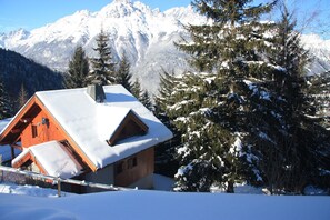 A wooden cabin with a sloped roof covered in snow, surrounded by snow-covered trees and mountains in the background.