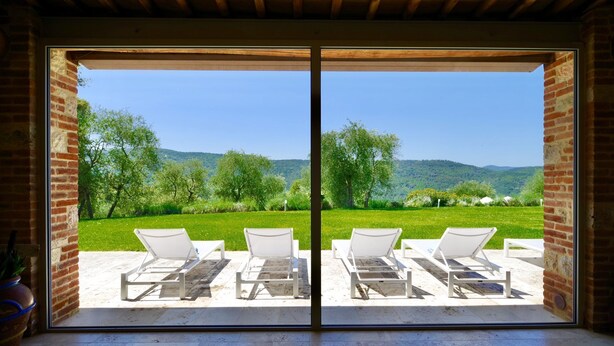 Une pièce dotée d’une grande porte-fenêtre coulissante qui donne sur un espace extérieur aménagé avec deux fauteuils de jardin blancs et offrant une vue sur un paysage verdoyant, avec des arbres et des collines.