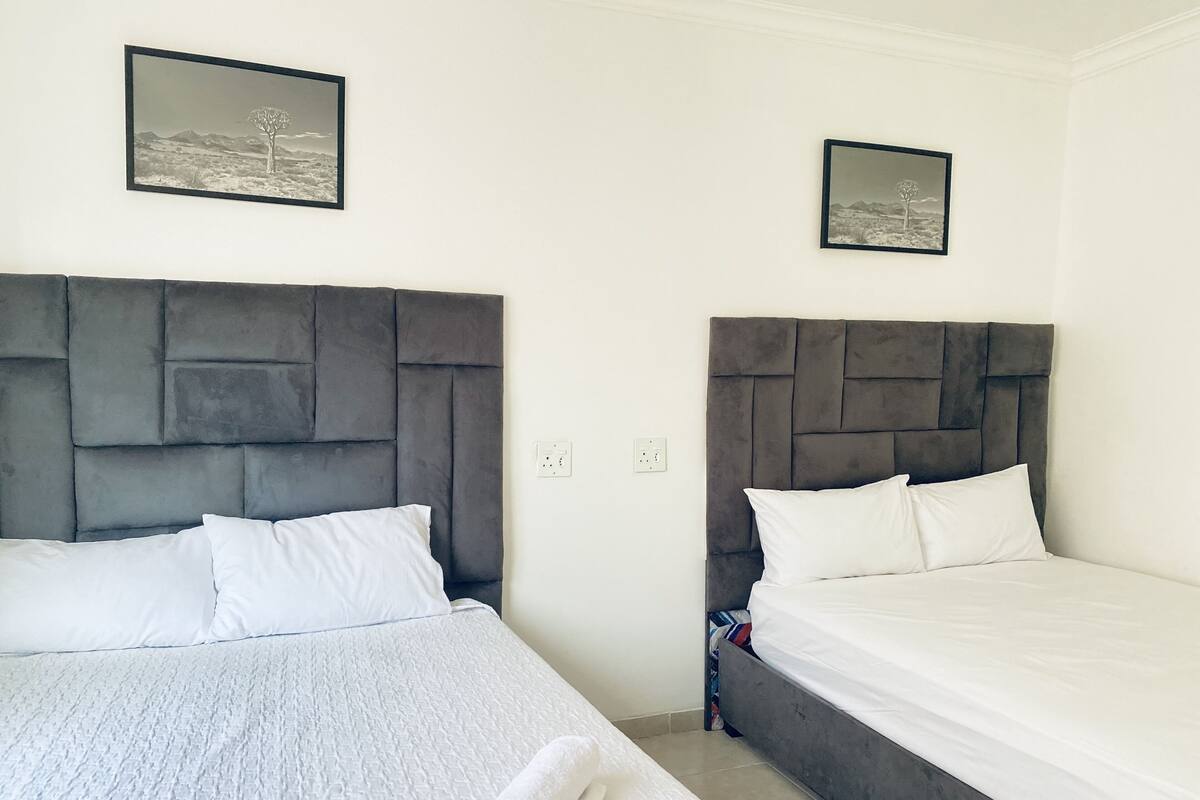 Une chambre d’hôtel avec deux lits, chacun doté d’une tête de lit rembourrée de couleur grise et de linge de lit blanc. Deux tableaux encadrés sont accrochés au mur au-dessus des lits.