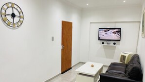 A living room with a white wall, a wooden door, a white coffee table, a black sofa, a TV mounted on the wall, and a ceiling fan. - Airport Royal Guest House (Kempton Park)