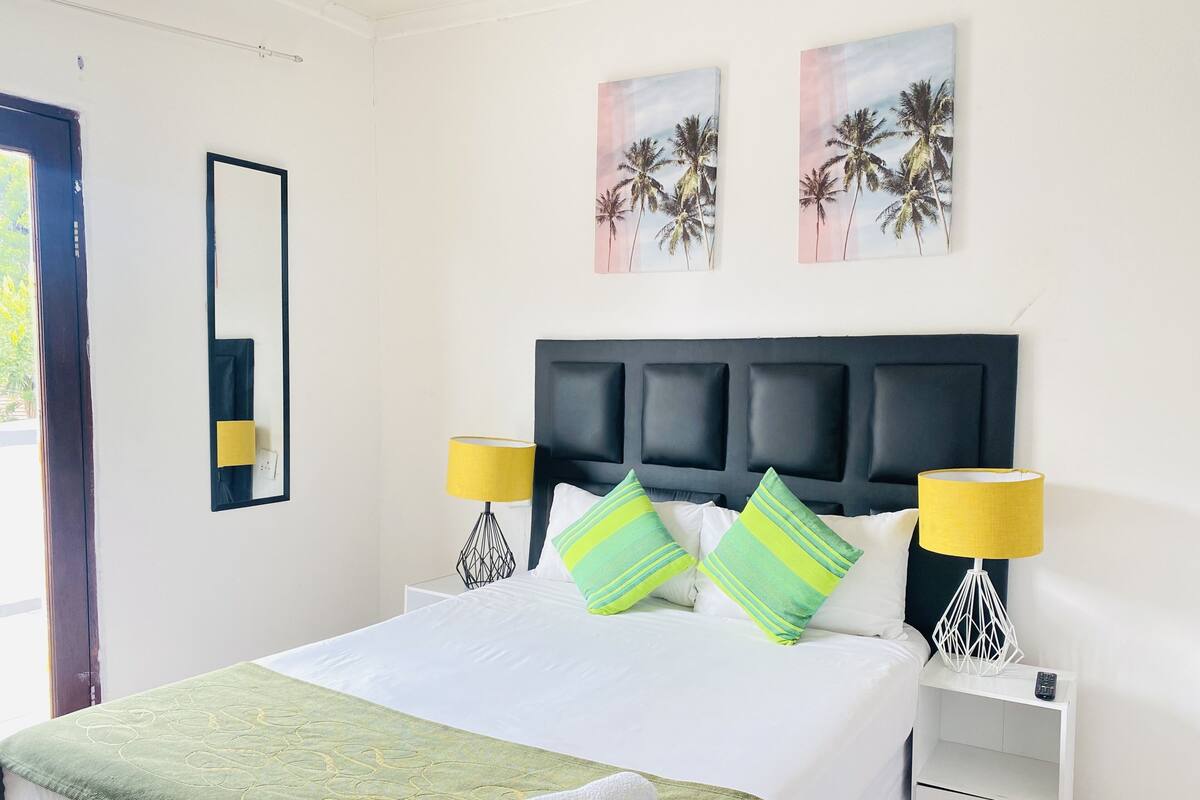Un lit bien fait, recouvert d’une couverture verte, avec deux oreillers et une couverture blanche. On y trouve deux tables de chevet avec des lampes, un miroir et des œuvres d’art accrochées au mur.