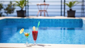 Ein Poolbereich mit zwei Cocktails auf einem Tisch, einem Rettungsschwimmerstuhl und Topfpflanzen im Hintergrund.