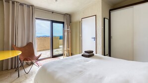 Quarto de hotel com cama, poltrona, mesa, porta de vidro deslizante para uma varanda e vista para o exterior.