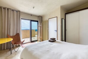 A hotel room with a bed, a chair, a table, a sliding glass door to a balcony, and a view of the outdoors.