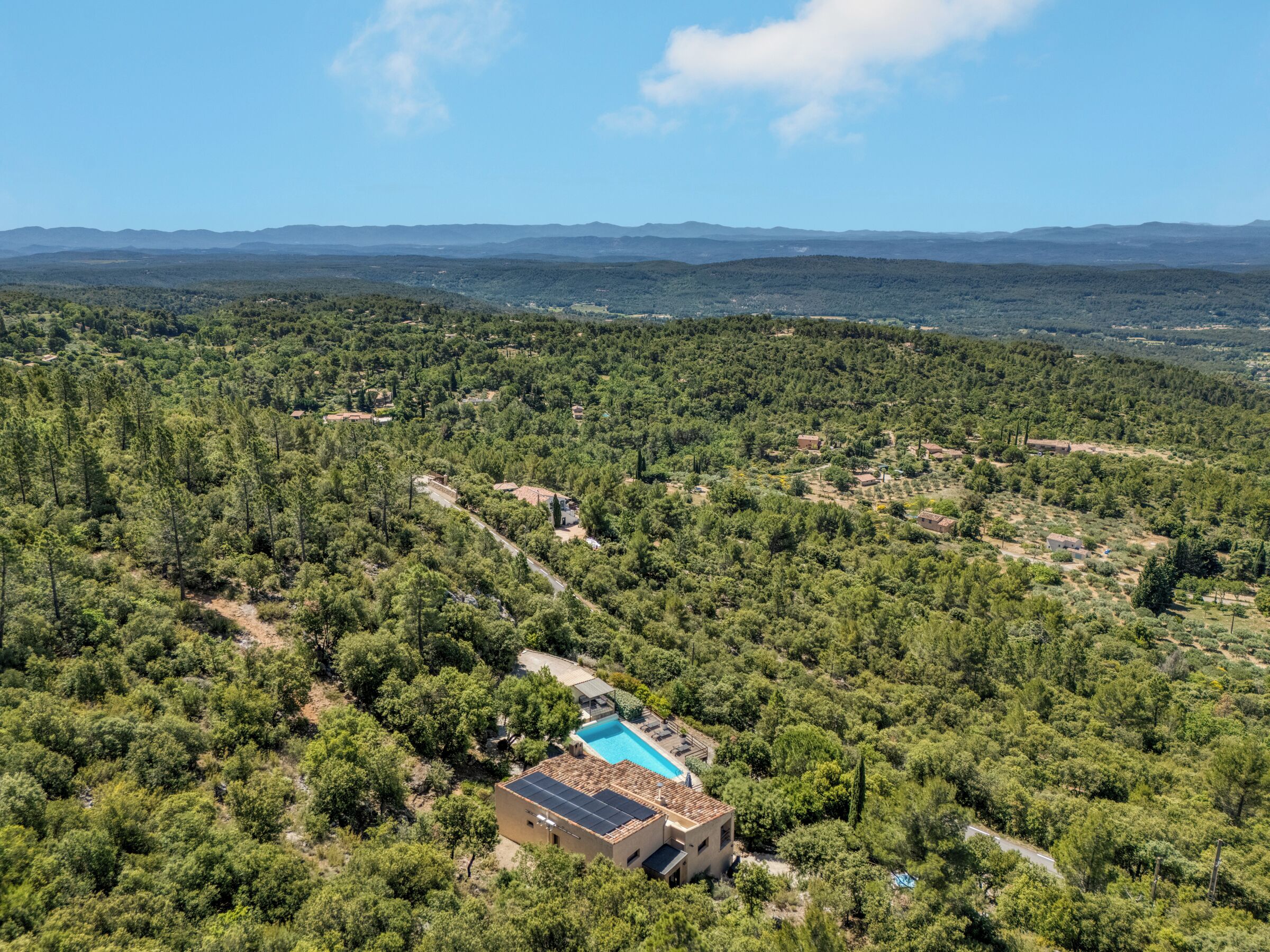 An aerial view of a property with a swimming pool, surrounded by dense forest and distant mountains.