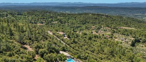 An aerial view of a property with a swimming pool, surrounded by dense forest and distant mountains.