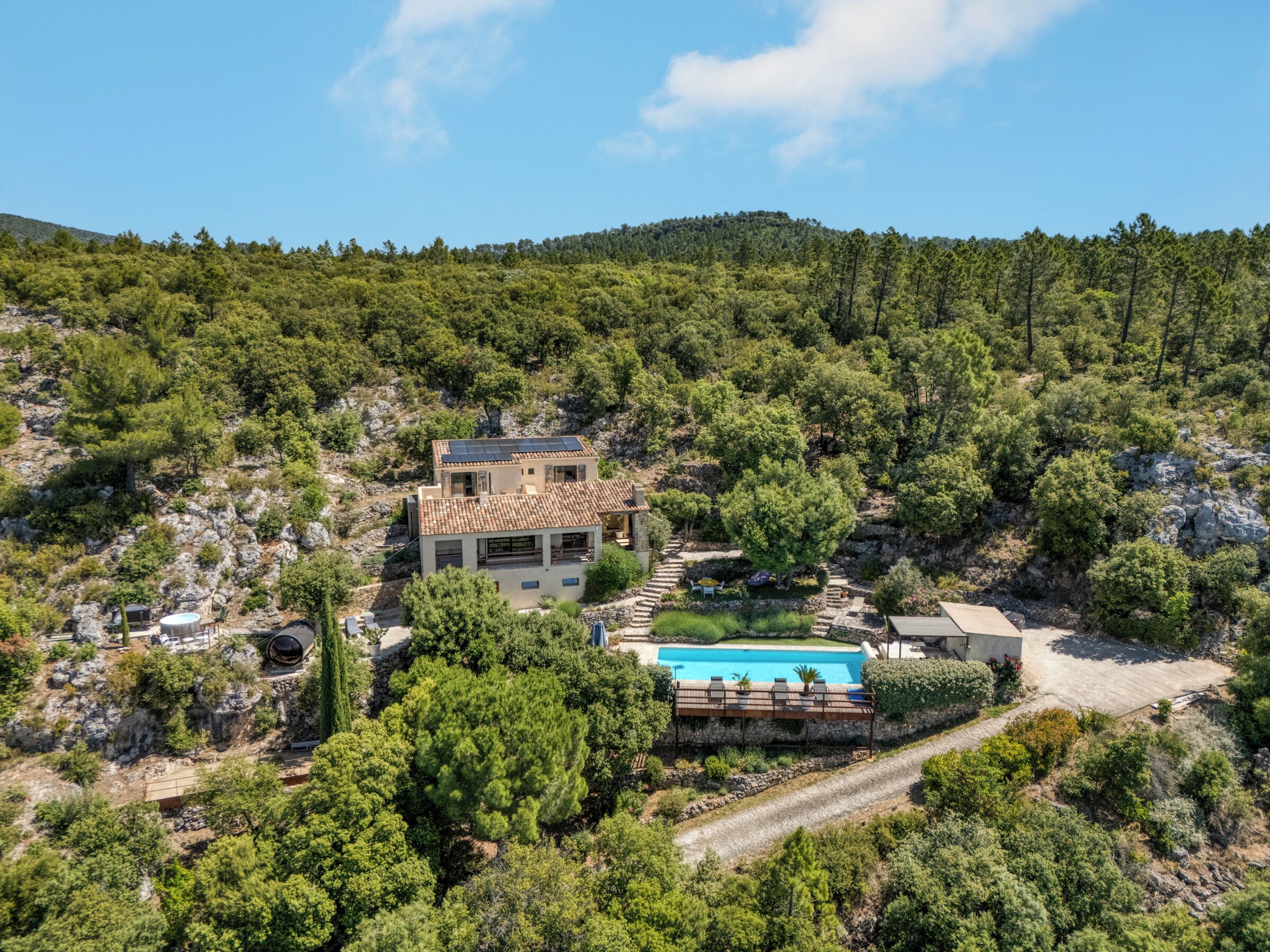 Uma casa de dois andares com piscina, rodeada por uma floresta densa e terreno rochoso.