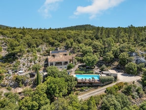 A two-story house with a swimming pool, surrounded by dense forest and rocky terrain.