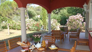 Un espace extérieur abrité, aménagé pour les repas, avec des meubles en bois, une table dressée avec du pain, des pâtisseries et des boissons, le tout entouré de fleurs épanouies et de verdure.