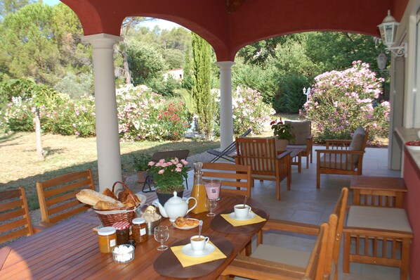 Un espace extérieur abrité, aménagé pour les repas, avec des meubles en bois, une table dressée avec du pain, des pâtisseries et des boissons, le tout entouré de fleurs épanouies et de verdure.