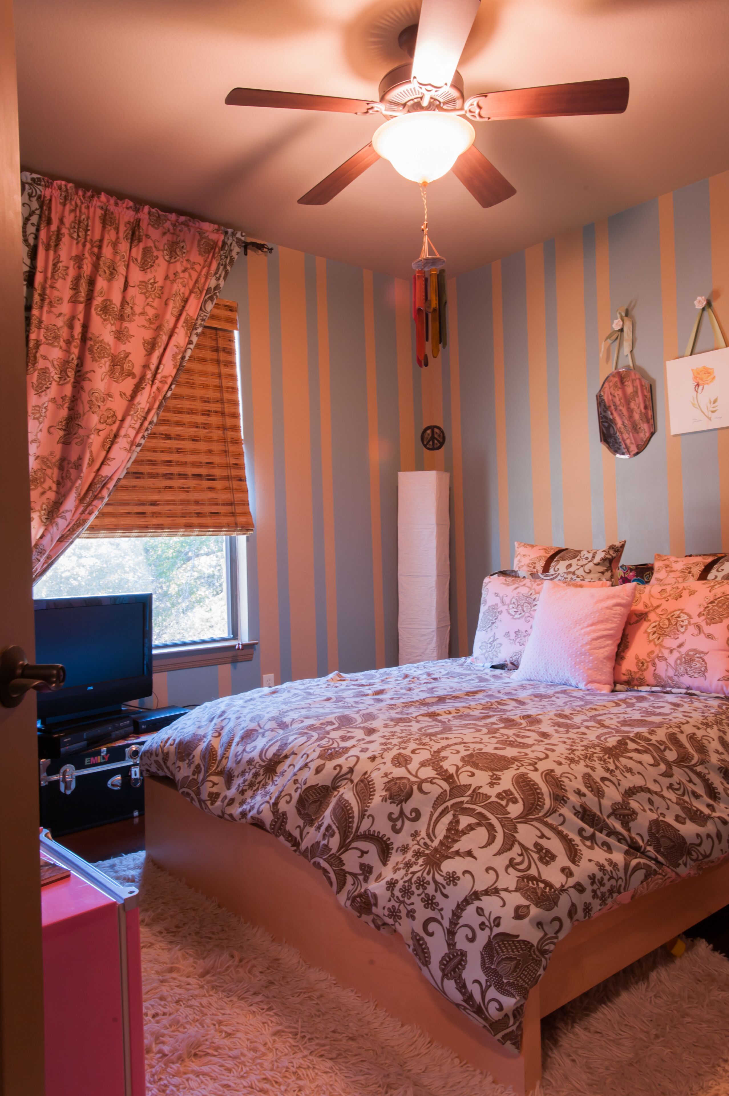 A bedroom with a bed, a ceiling fan, a window with curtains, a dresser, and a wall with vertical stripes.