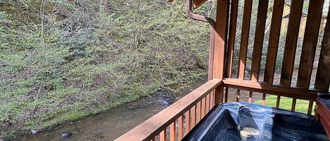 A wooden deck with a hot tub overlooking a stream and dense forest.