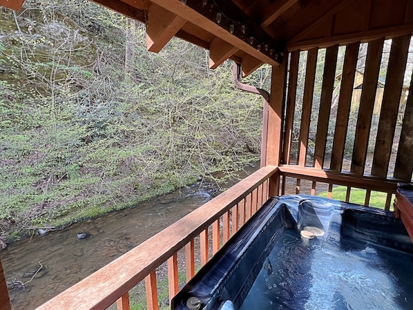 Eine Holzterrasse mit Whirlpool, mit Blick auf einen Bach und einen dichten Wald.